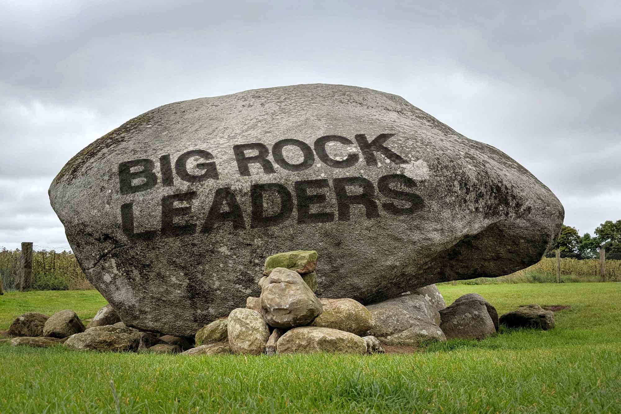The Big Rock Leaders name chiseled in bold capital letters across the face of a huge grey glacial boulder resting on smaller support stones in a green field under a pale overcast sky