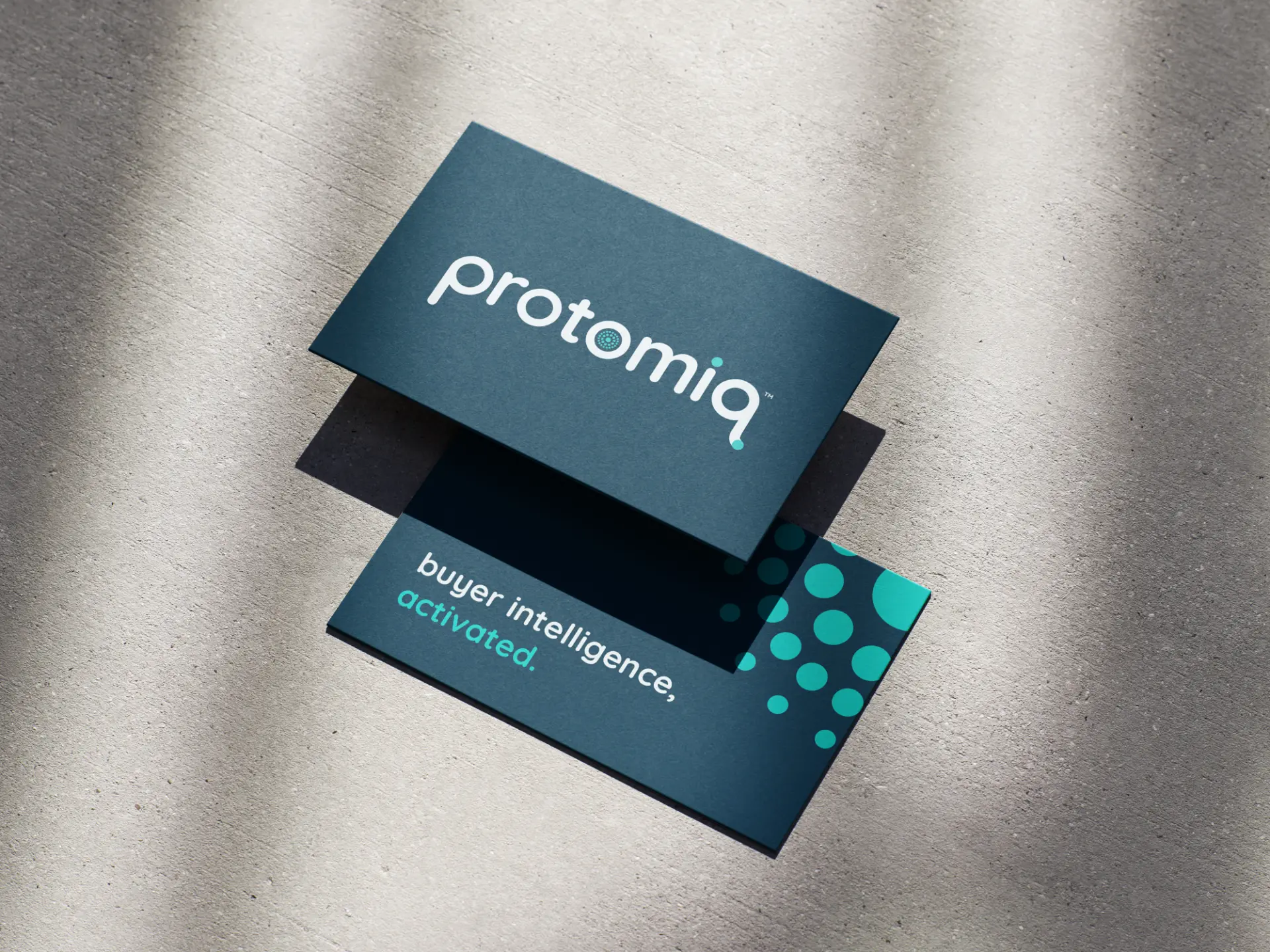 Two Protomiq business cards on a concrete surface, showing the lowercase protomiq wordmark and the line buyer intelligence, activated